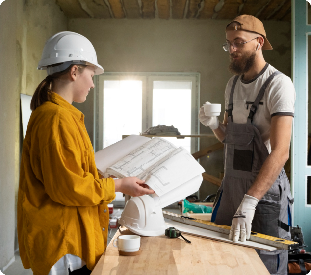 Female contractor in hard hat reviewing construction plans with male worker holding coffee cup at renovation site