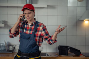 A repair technician wearing a red cap and checkered shirt talks on the phone in a kitchen, looking confused while holding a metal tool in one hand.